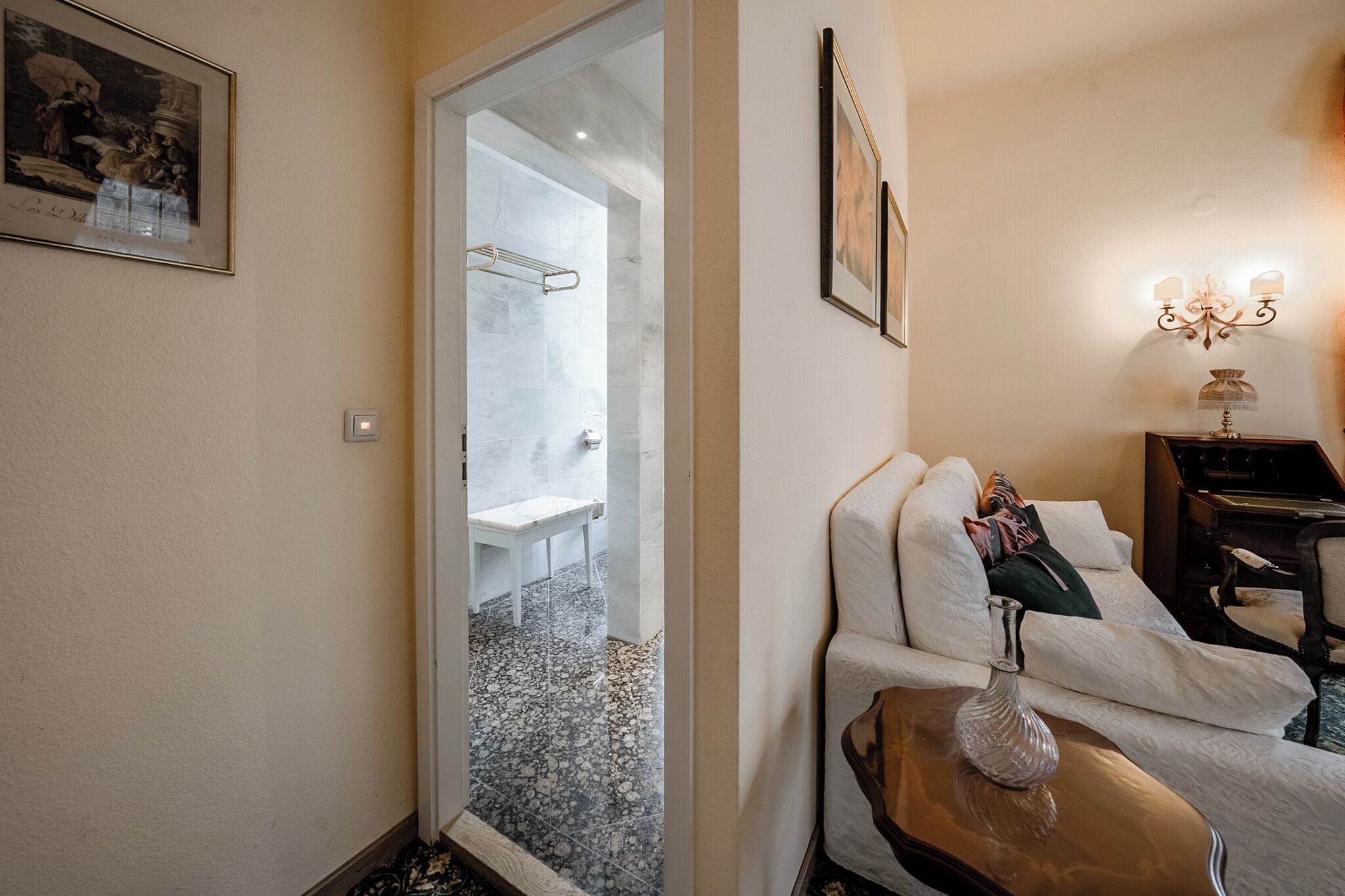 Une chambre d’hôtel avec une porte en verre donnant sur la salle de bain, un canapé blanc avec des coussins, une petite table d’appoint en bois et un luminaire fixé au mur.