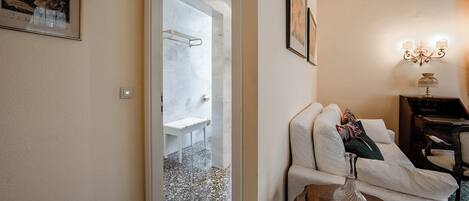 Une chambre d’hôtel avec une porte en verre donnant sur la salle de bain, un canapé blanc avec des coussins, une petite table d’appoint en bois et un luminaire fixé au mur.