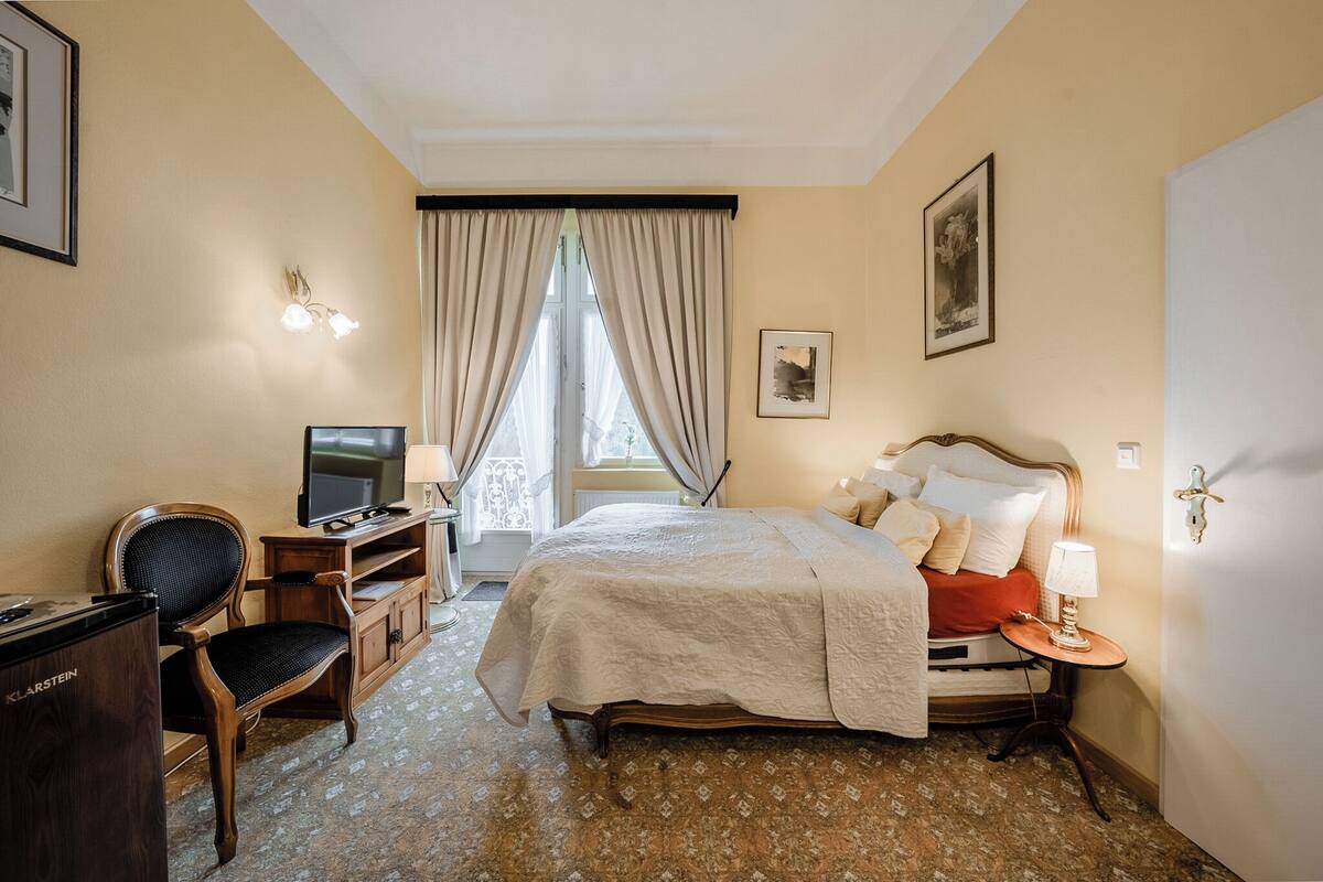A hotel room with a bed, a desk with a television, a chair, and a small table with a lamp.