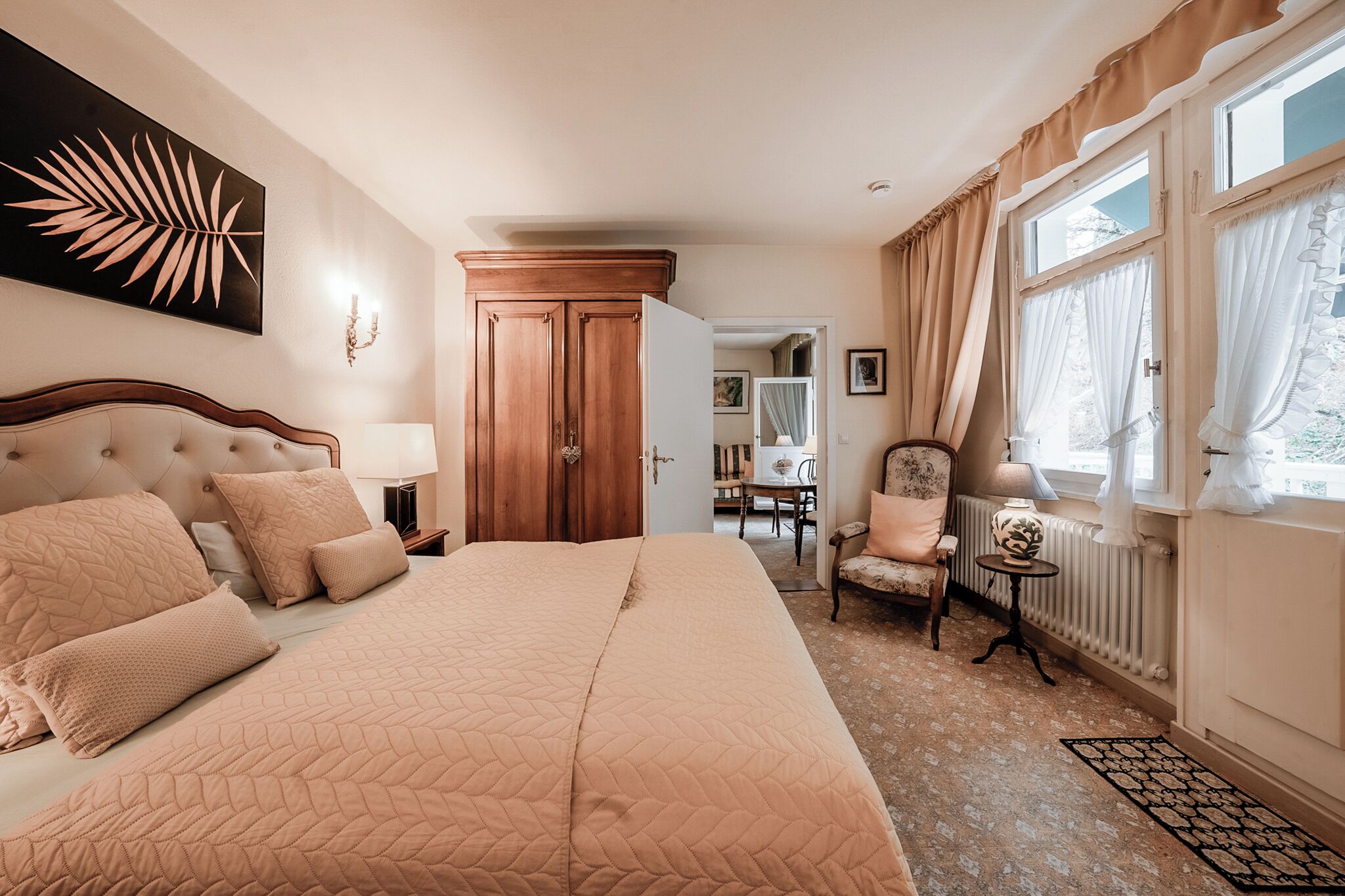 A bedroom with a large bed, a wooden wardrobe, a chair, a small table, and a window with curtains.