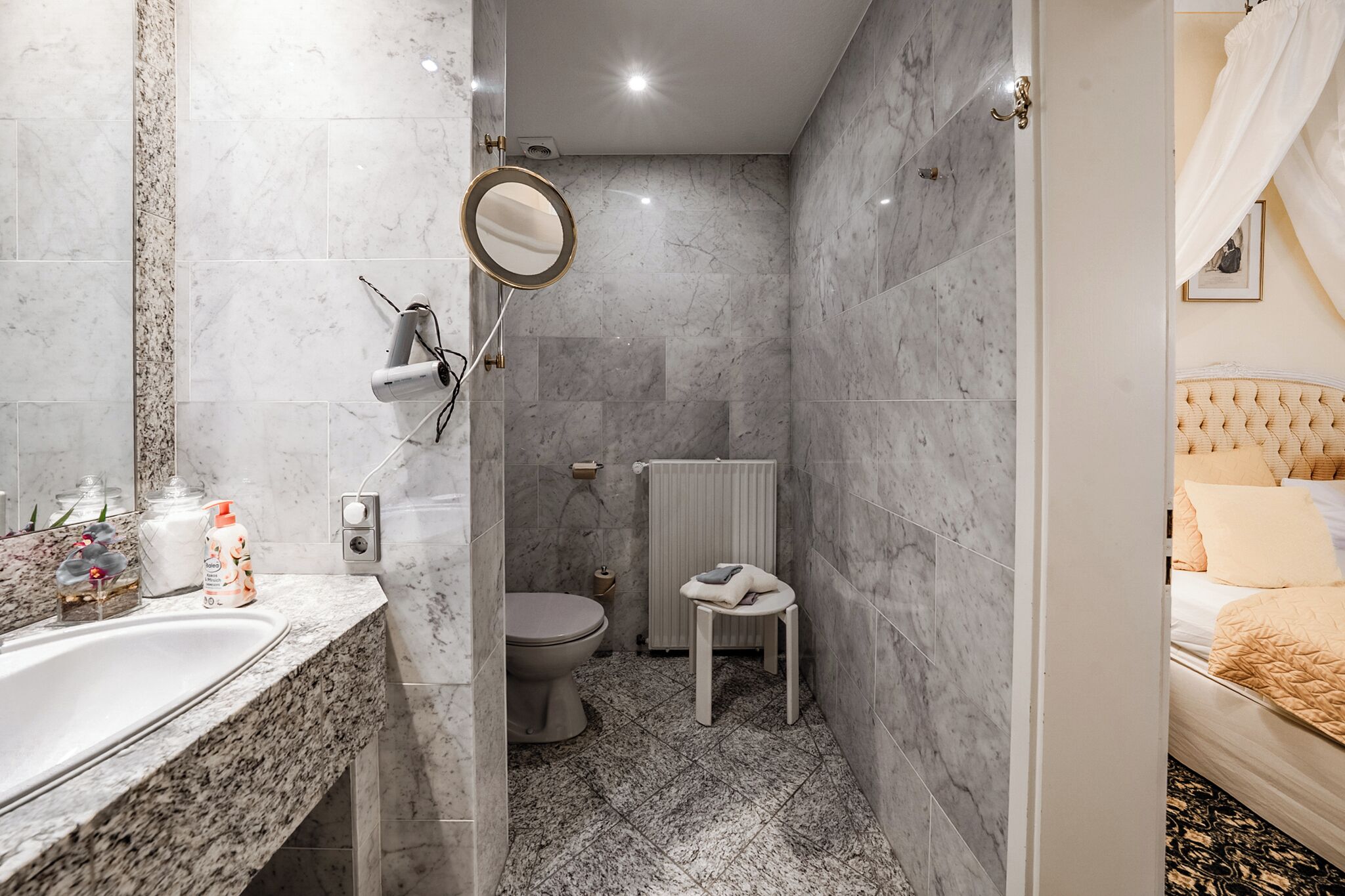 A compact bathroom with a marble floor, a toilet, a sink with toiletries, and a bedside table with a pillow.