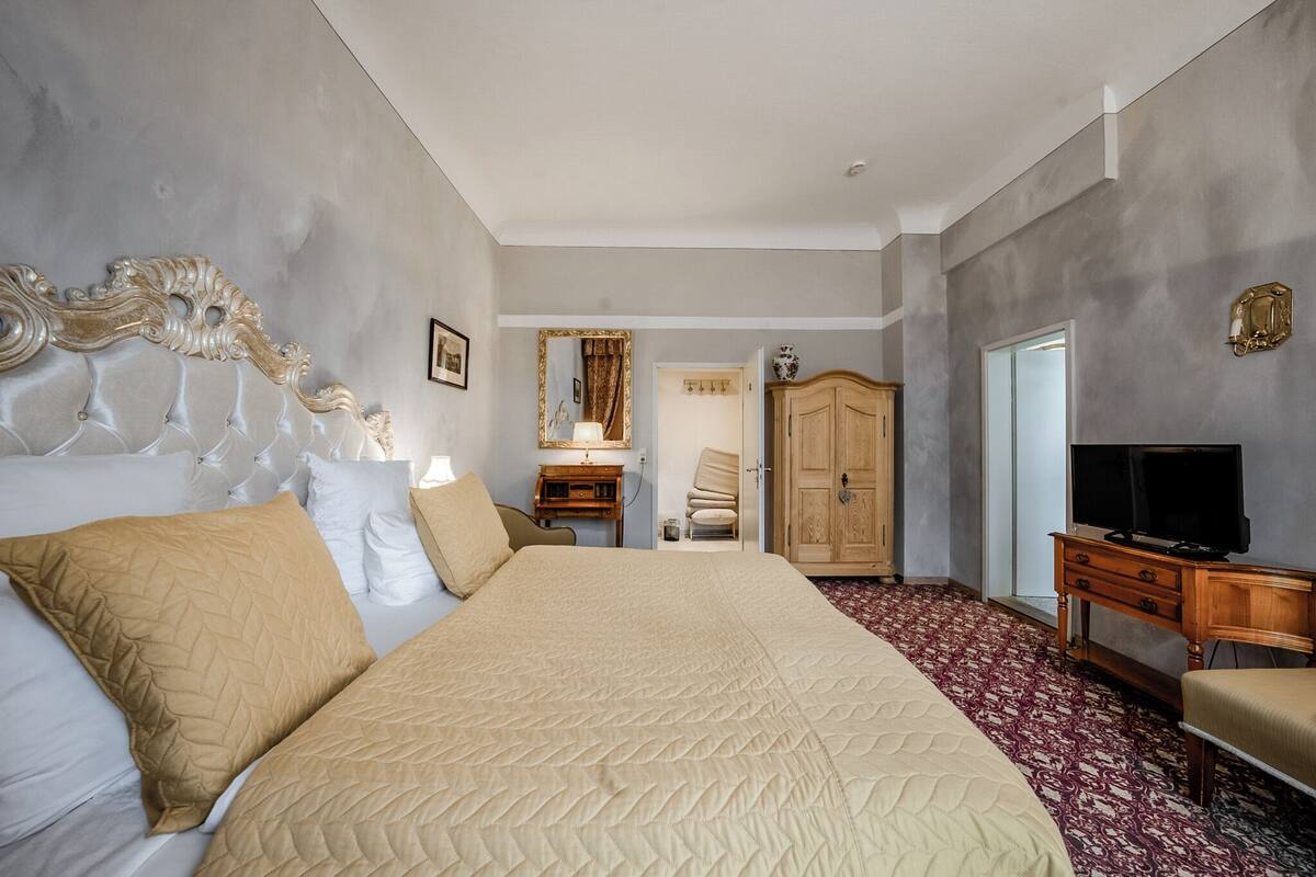 A hotel room with a large bed, a wooden wardrobe, a television, and a desk with a lamp.