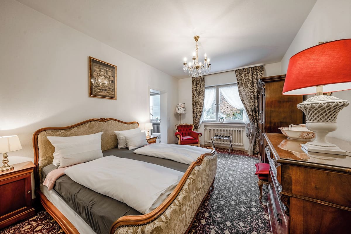 A bedroom with a large bed, a chandelier, a red chair, and a wooden dresser.
