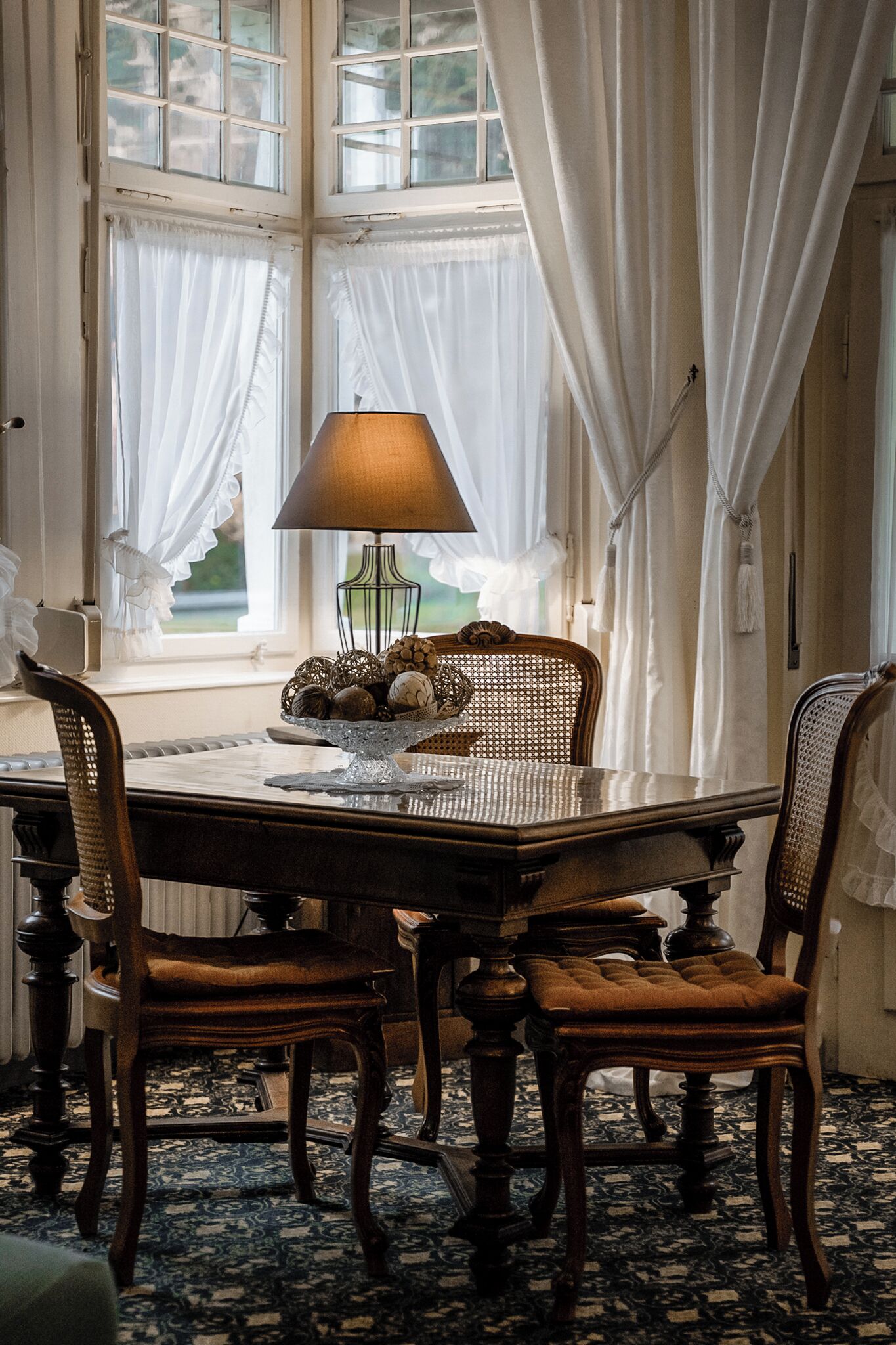 A dining area with a wooden table, four chairs, a lamp, and a window with curtains.