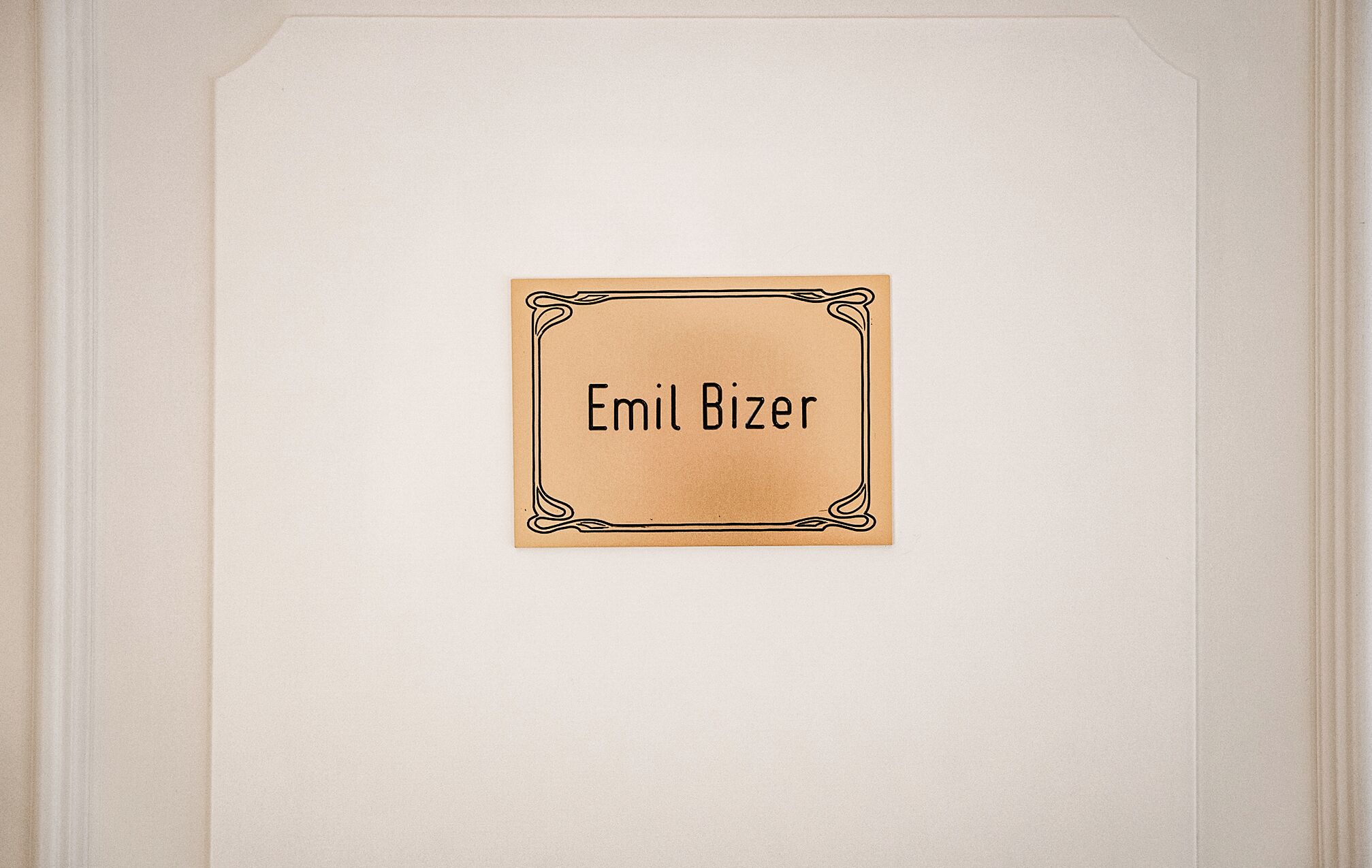 A framed nameplate with "Emil Bizer" on a plain wall.