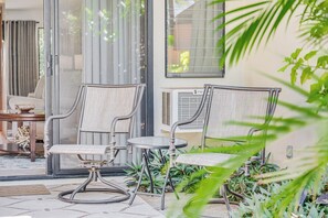 Eine Terrassenfl äche mit zwei Korbstühlen und einem kleinen Tisch, umgeben von Blumentöpfen und einer Glastür, die zu einem Raum führt.