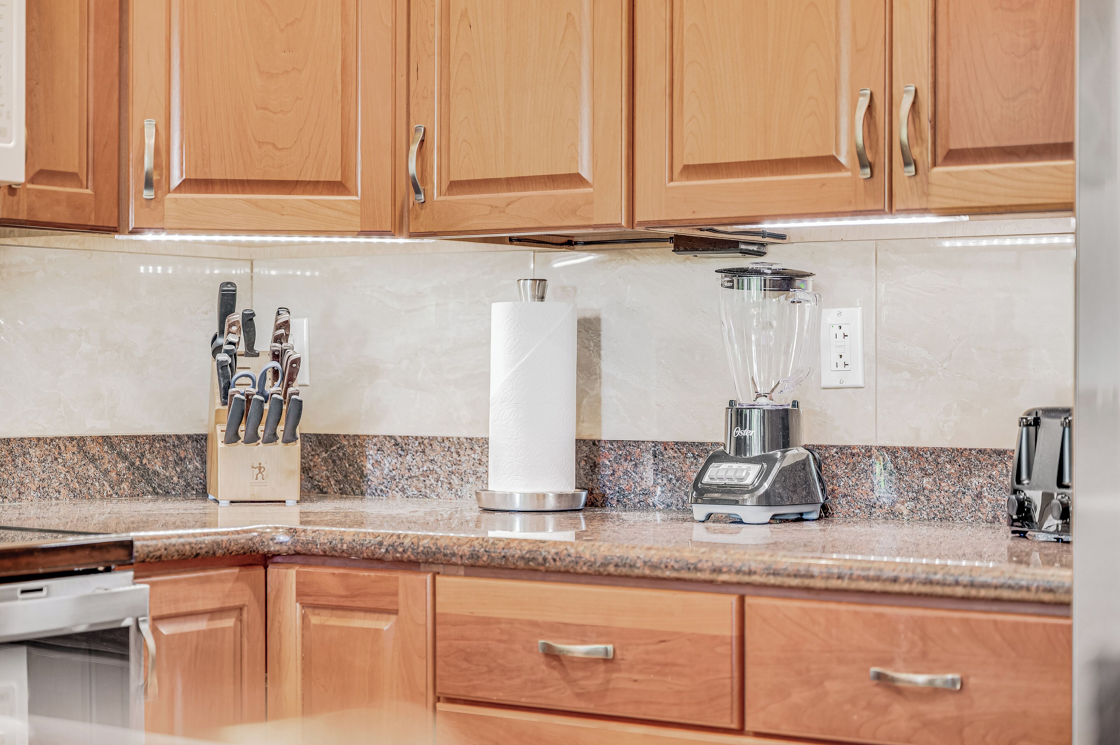A kitchen with wooden cabinets, a granite countertop, a knife block, a paper towel holder, a blender, and a toaster.