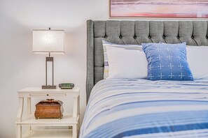 A neatly made bed with a striped blue and white comforter, a grey upholstered headboard, a bedside lamp, and a small wooden chest. - Maui Kamaole #I-106 Ground Flr, Garden View, A/C, Great Location (Kihei)