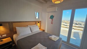 Una camera da letto con un letto grande, una testiera in legno, un condizionatore d'aria, un comodino e vista sul paesaggio costiero attraverso una grande finestra.