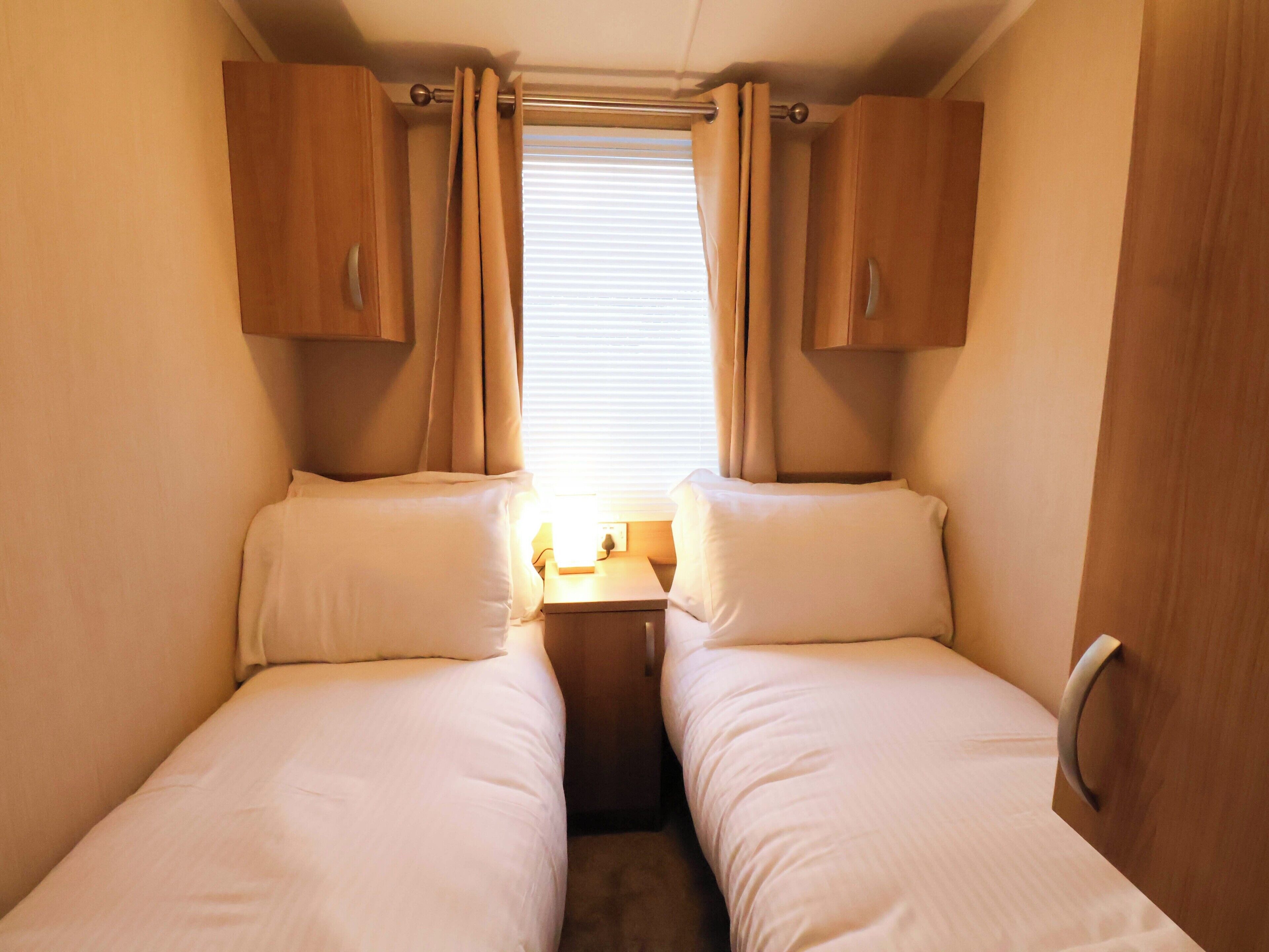 A small, cozy room with two beds, wooden cabinets, a window with curtains, and a small table with a lamp.