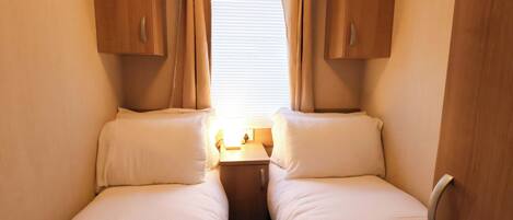 A small, cozy room with two beds, wooden cabinets, a window with curtains, and a small table with a lamp.