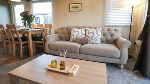Een woonkamer met een beige bank, een houten bijzettafeltje met kaarsen en een boek, een eethoek met een tafel en stoelen, en een ingelijst beeld aan de muur.