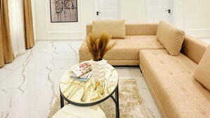 Een moderne woonkamer met een beige modulaire bank, een ronde salontafel met een vaas en boeken, een marmeren vloer, en een kunstwerk in een lijst aan de muur.