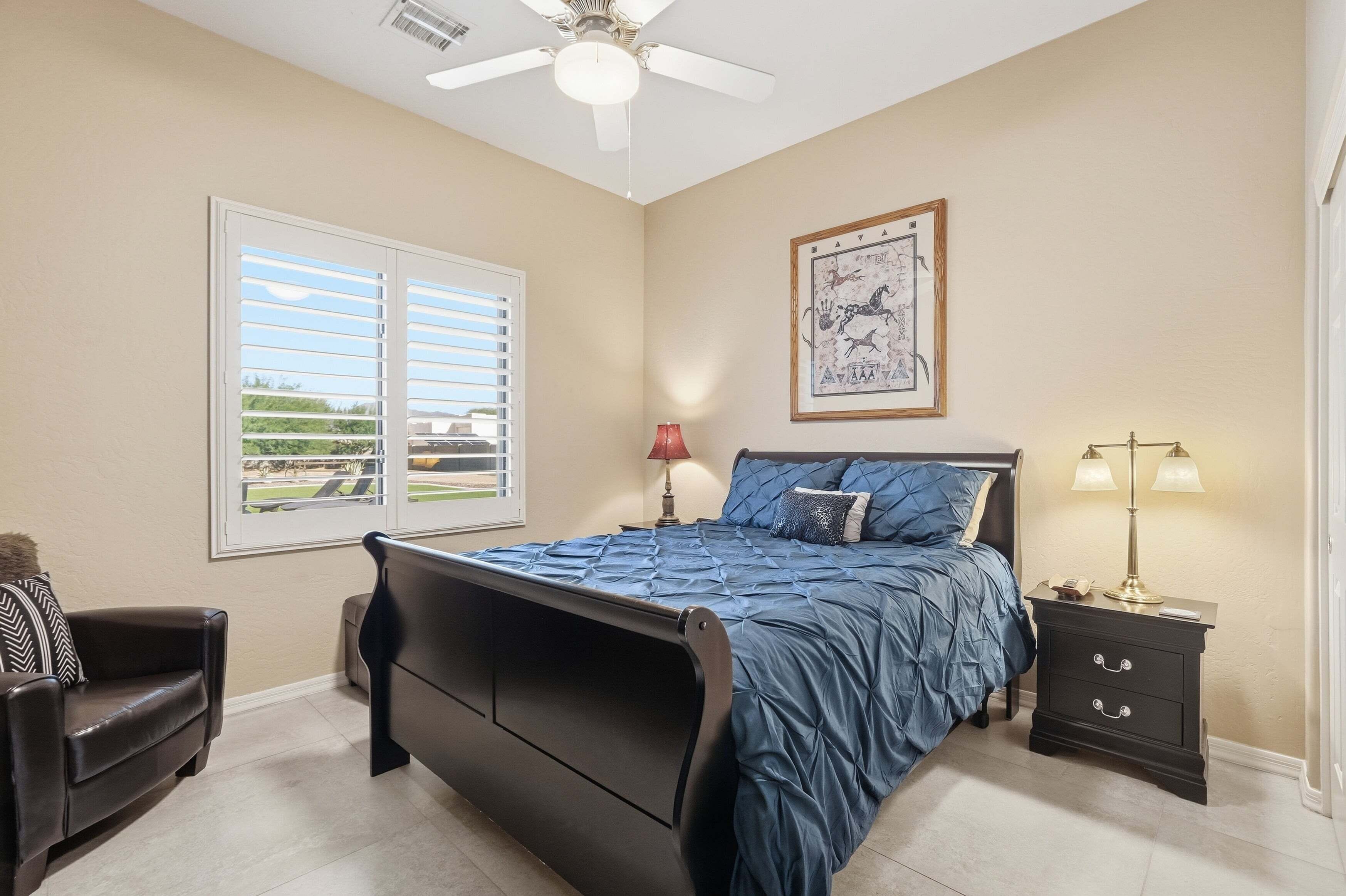 A bedroom with a bed, a nightstand, a chair, a window with blinds, and a ceiling fan.