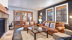 A living room with a stone fireplace, leather sofas, a glass coffee table, and large windows with blinds. - Town Pointe A105 3 Bedroom Condo (Park City)