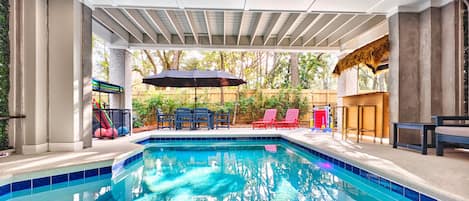 Una piscina con borde de azulejos, rodeada por un porche cubierto con asientos y una estructura de juegos al fondo.