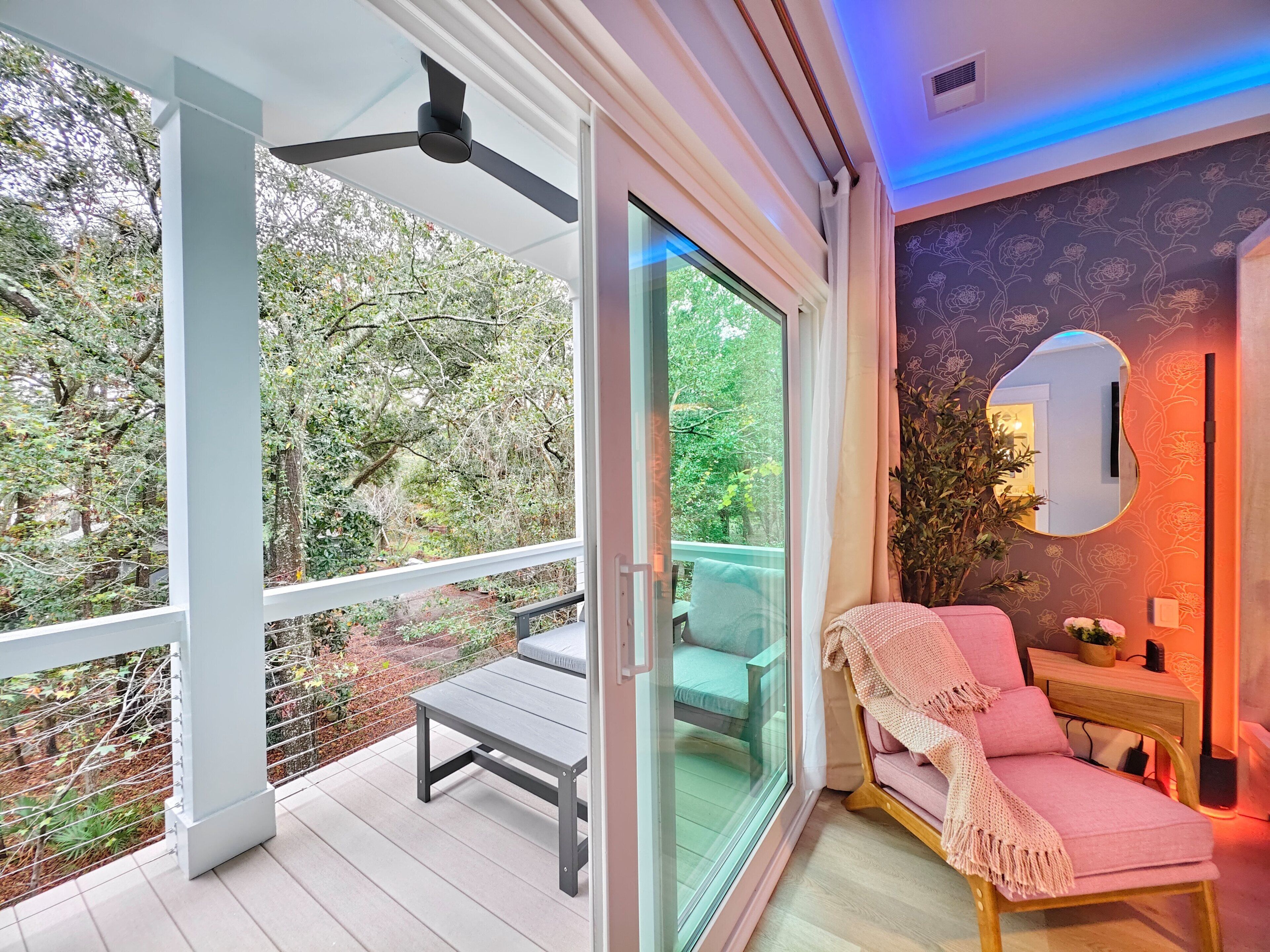 A modern room with a sliding glass door leading to a balcony, a ceiling fan, a pink chair, and a wall-mounted mirror.