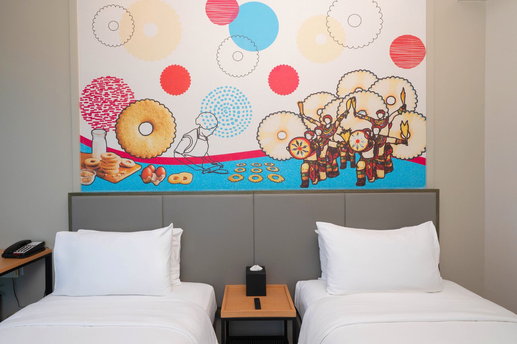 Ein Hotelzimmer mit zwei Betten, einem Nachttisch mit Telefon und Taschentuchbox sowie einem farbenfrohen Wandbild, das Donuts, eine Person und eine Szene aus der Kultur der amerikanischen Ureinwohner zeigt.