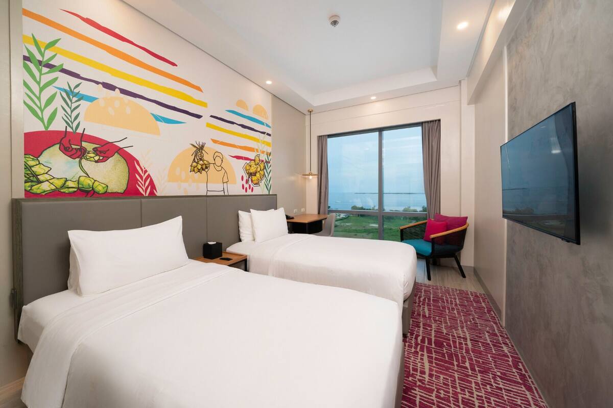 Ein Hotelzimmer mit zwei Betten, einem großen Fenster mit Aussicht, einem Fernseher und einem farbenfrohen Wandgemälde.