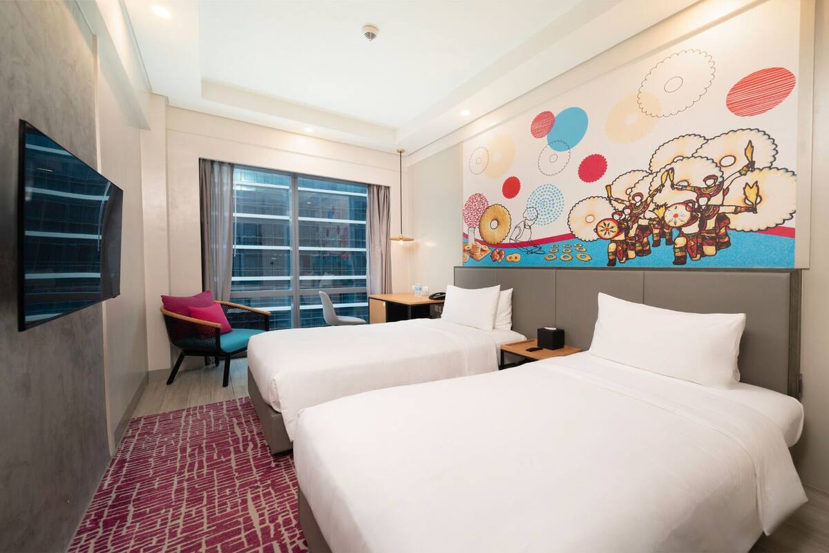 Ein Hotelzimmer mit zwei Betten, einem Fernseher, einem großen Fenster und einem farbenfrohen Wandgemälde.