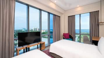 Habitación de hotel con dos camas, un televisor sobre un soporte de madera, un escritorio y ventanas amplias con vistas a la ciudad.