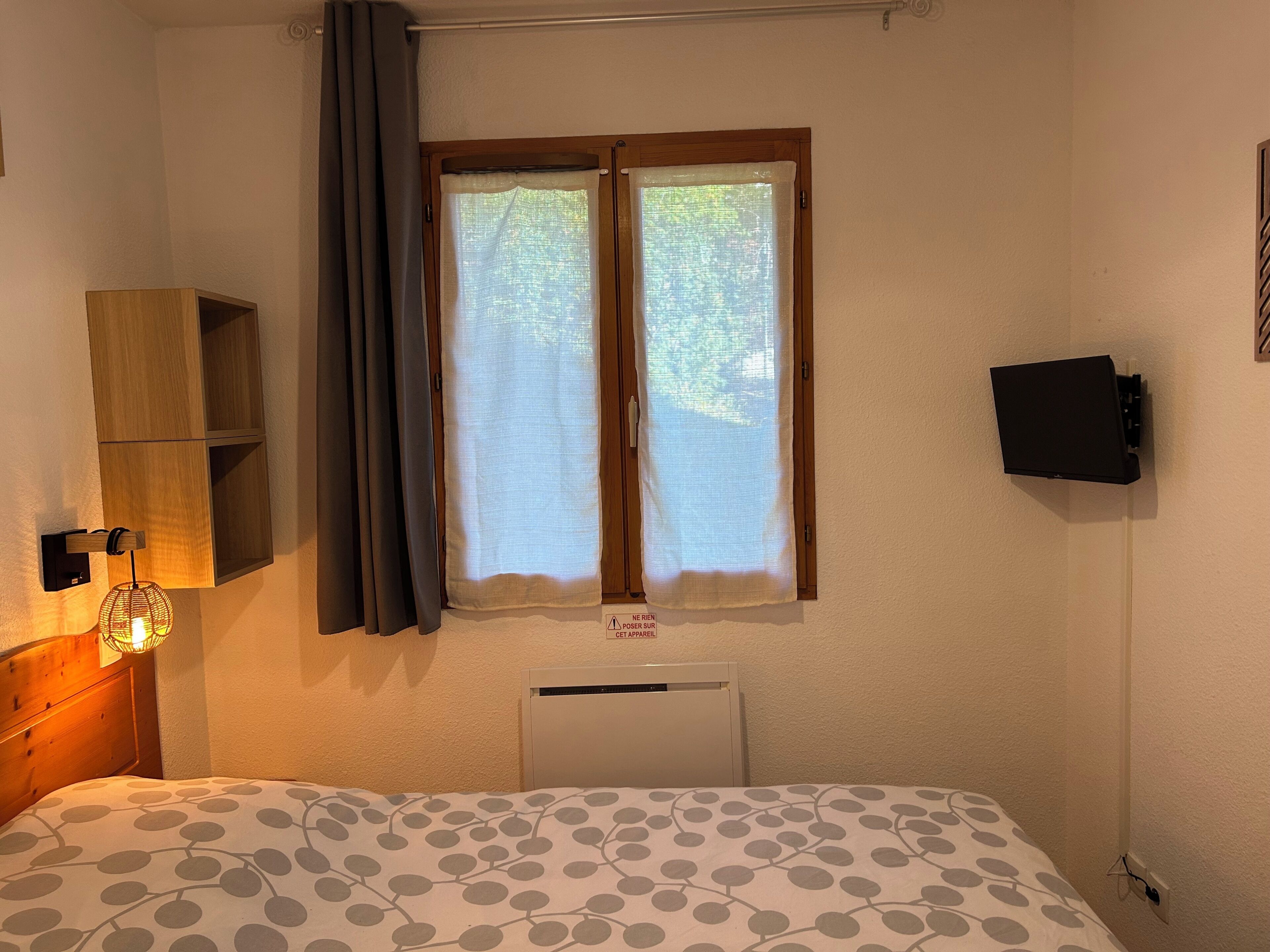 Une chambre à coucher avec un lit, une fenêtre avec des rideaux, un téléviseur fixé au mur et un radiateur.