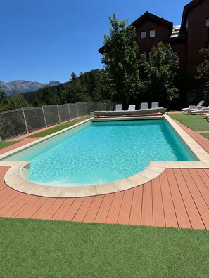 Ein rechteckiger Pool mit umlaufender Holzterrasse, einer Rasenfläche und einem Gebäude mit Satteldach im Hintergrund.