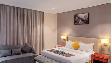 Habitación de hotel con cama, mesitas de noche, un sofá y un amplio ventanal con cortinas.