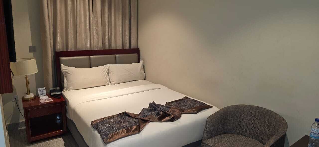 A hotel room with a bed, a chair, a nightstand with a lamp, and a water bottle.