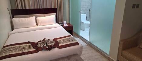 A hotel room with a large bed, a nightstand, a bathroom with a toilet and shower, and a chair.