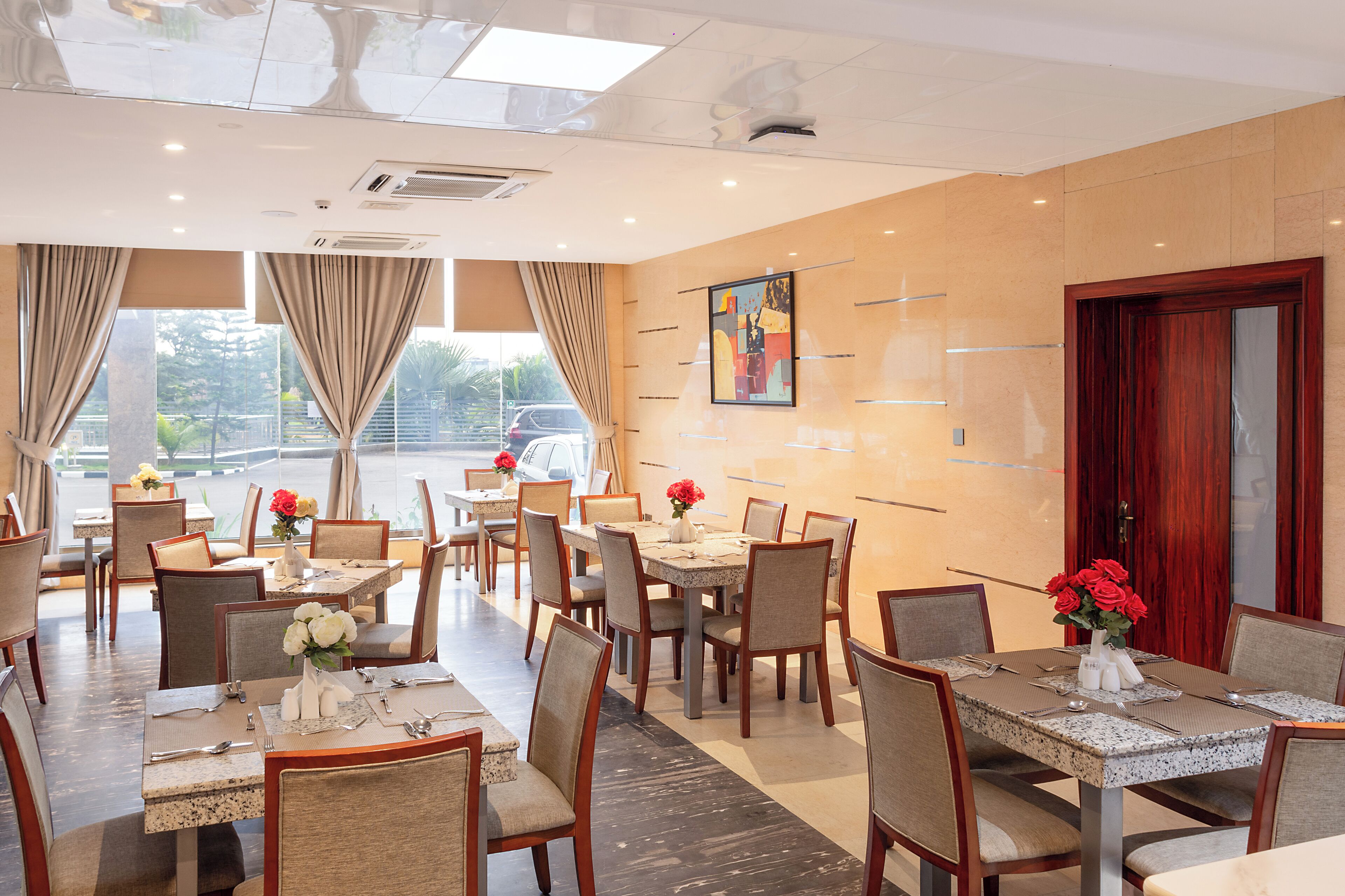 A hotel dining area with multiple tables and chairs, large windows with curtains, and a painting on the wall.