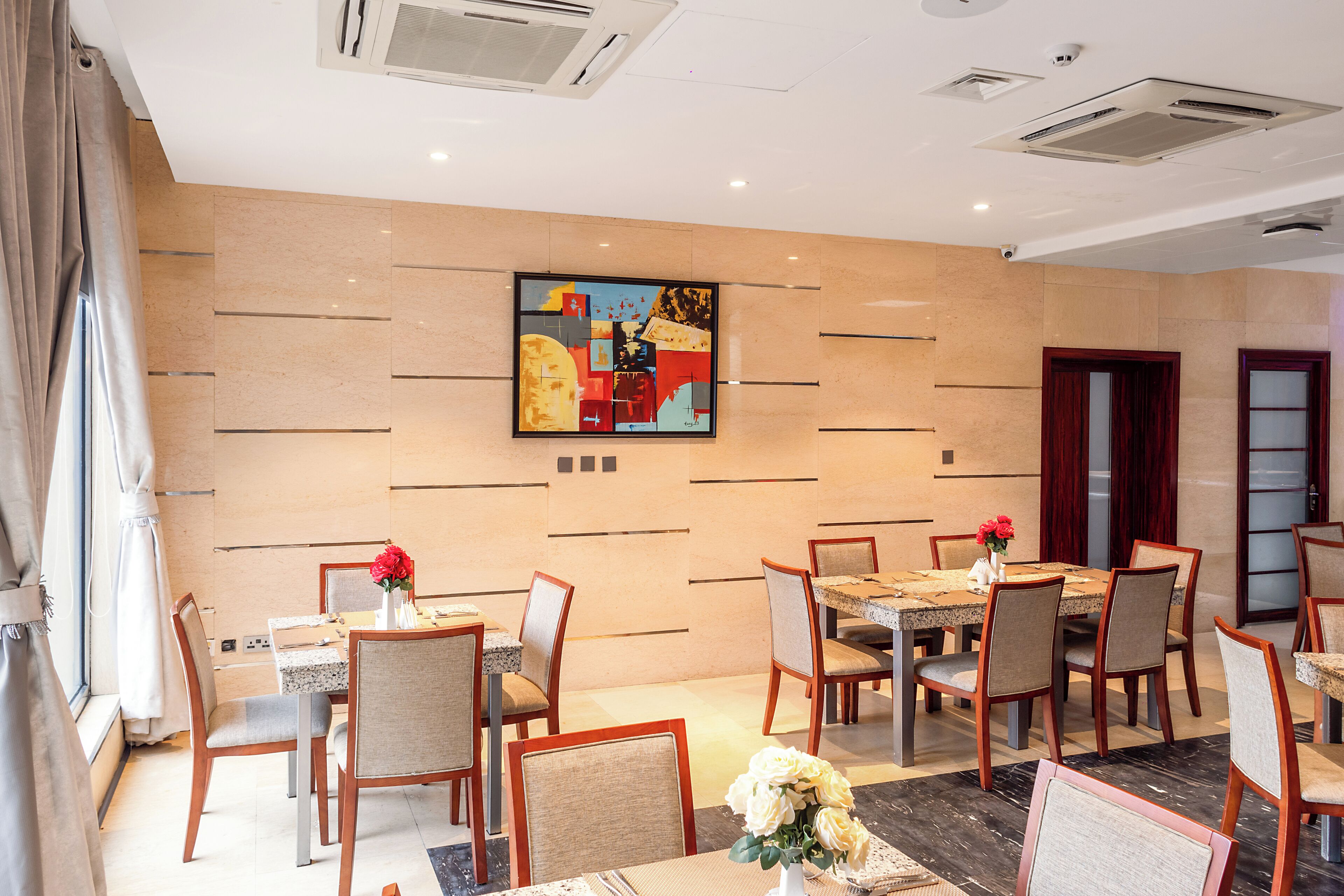 A hotel dining area with multiple tables and chairs, a large abstract painting on the wall, and a modern ceiling design.