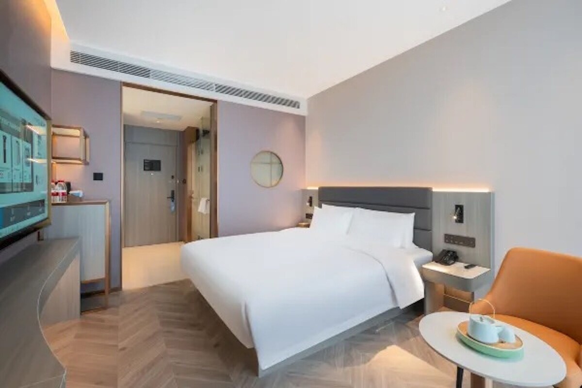 A hotel room with a bed, bedside table, chair, and a small table with a tray.