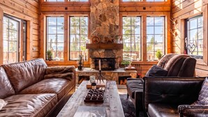A log cabin interior with a stone fireplace, leather sofas, and large windows.