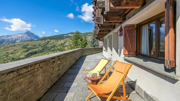 Un balcon avec deux fauteuils de jardin en bois, un mur en pierre et une vue sur la montagne.