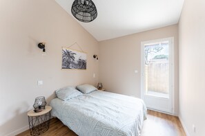 Un dormitorio con cama, una mesita de noche pequeña, una lámpara de pared, una obra de arte colgada y una ventana con vistas.