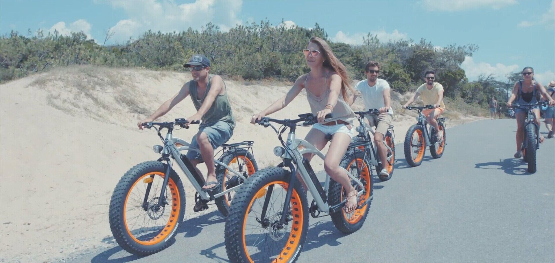 Een groep mensen fietst over een verharde weg, met een zandgebied aan één kant en begroeiing aan de andere kant.