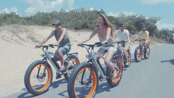 Een groep mensen fietst over een verharde weg, met een zandgebied aan één kant en begroeiing aan de andere kant.
