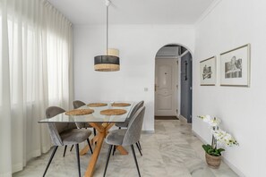 Een eetgedeelte met een glazen tafel, grijs gestoffeerde stoelen, een hanglamp en een doorgang naar een andere kamer.