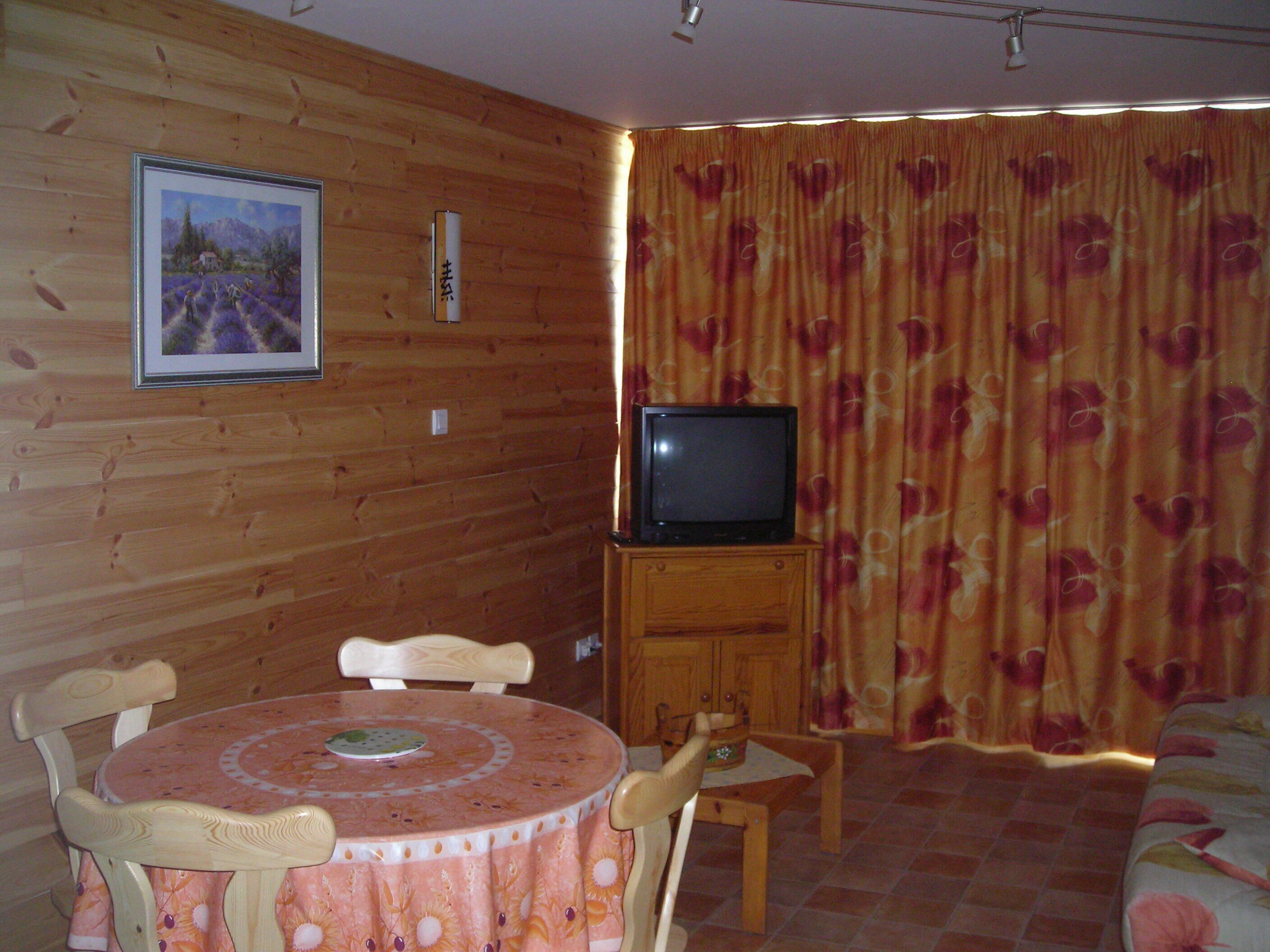 Ein Zimmer mit einer Holzwand, einem Fernseher auf einem Holztisch, einem Esstisch mit Stühlen und einem Bett mit floralem Überwurf.