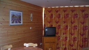 Une pièce avec un mur en bois, un téléviseur posé sur un meuble en bois, une table à manger avec des chaises et un lit recouvert d’une couvre-lit à motifs floraux.