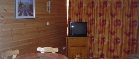 A room with a wooden wall, a television on a wooden stand, a dining table with chairs, and a bed with a floral bedspread.