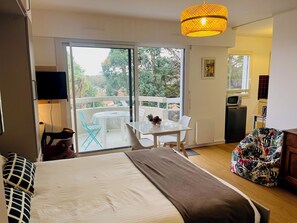 A bedroom with a bed, a small table with chairs, a sofa, a TV, and a balcony with a view of trees and houses. - Biarritz - Lahouze - Studio - Balcony - Parking Space   (Biarritz)