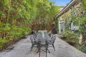 A covered patio with a table and chairs, surrounded by bamboo plants and a white building with a sign.