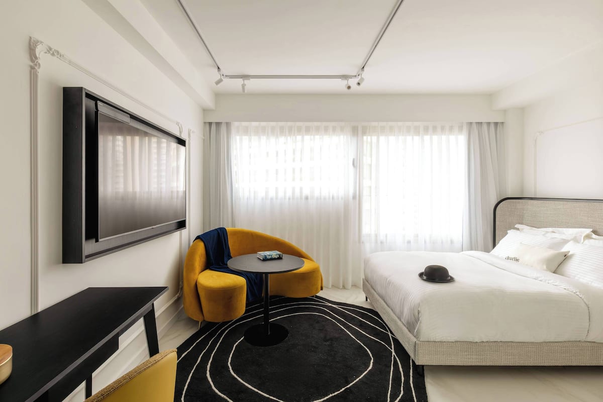 Una habitación de hotel moderna con televisión de pantalla plana, un sillón amarillo, una mesa redonda, una cama con ropa de cama blanca y una alfombra blanco y negro.