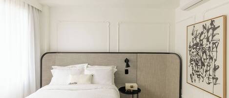 A hotel room with a large bed, a bedside table, a wall-mounted air conditioner, and a framed abstract painting.
