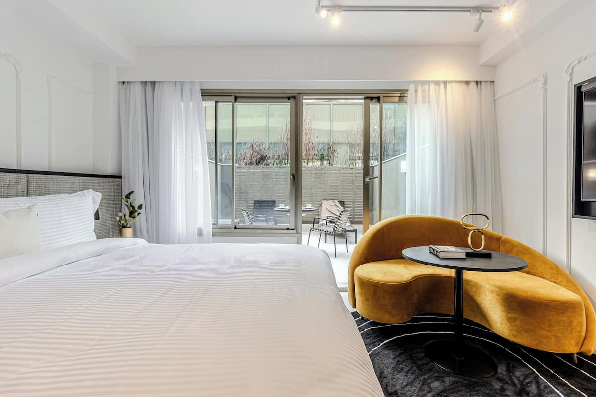 Una habitación de hotel con una cama grande, una mesita redonda, un sillón amarillo y un balcón con vistas.