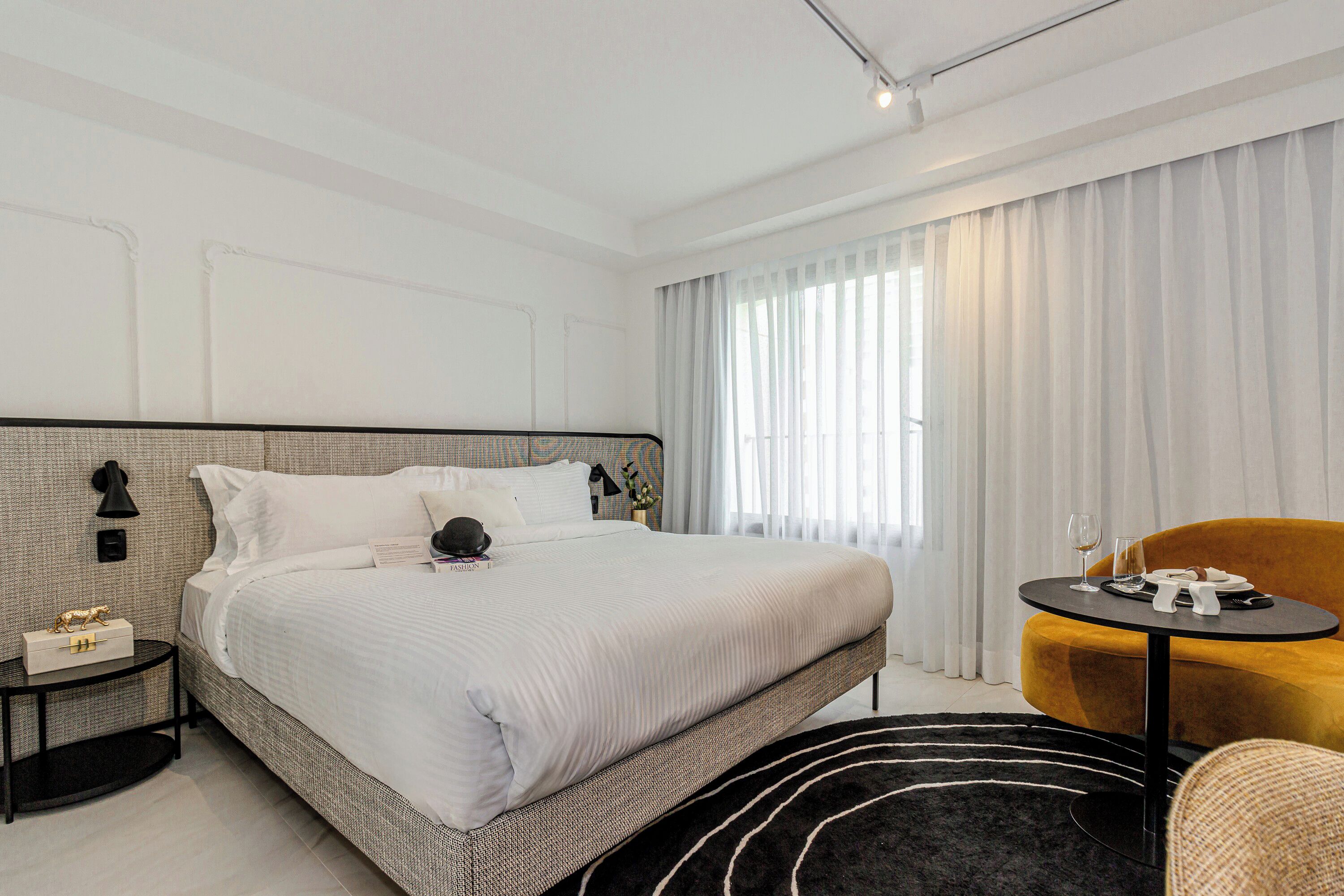 A modern hotel room with a large bed, a small side table, a round black table, and a yellow armchair.