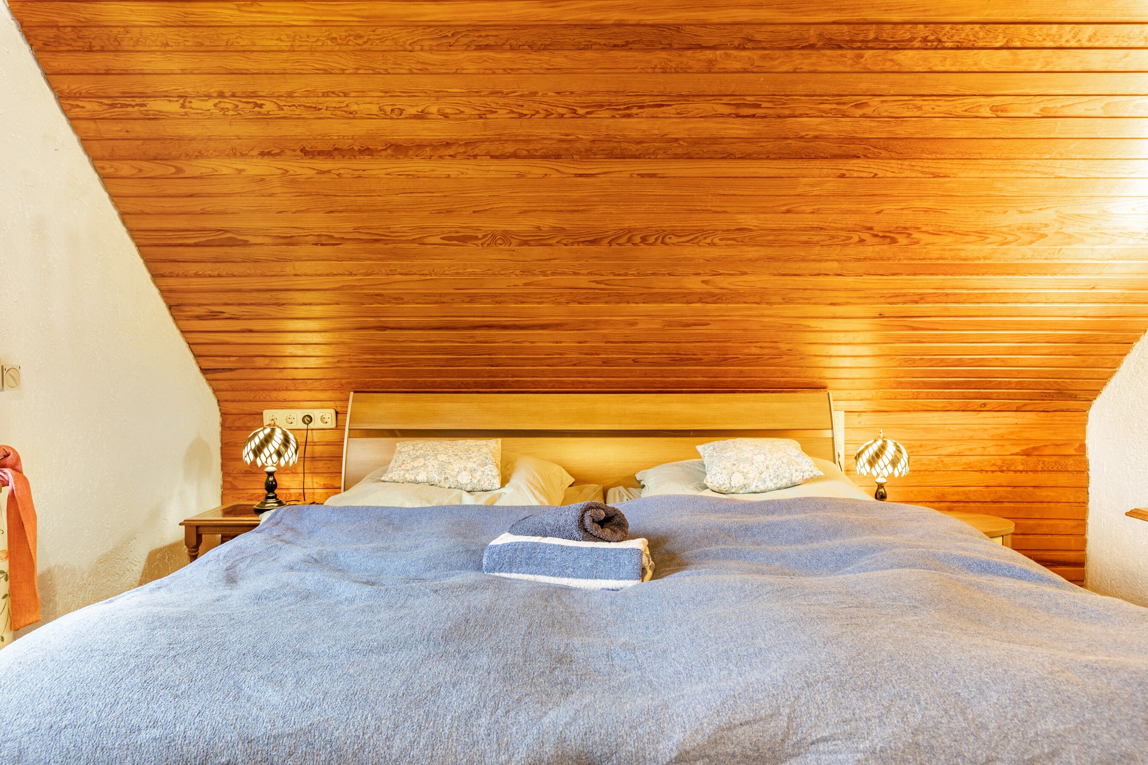 Une chambre à coucher avec un plafond en bois, un lit recouvert d’une couverture bleue, deux lampes de chevet et une table de nuit.