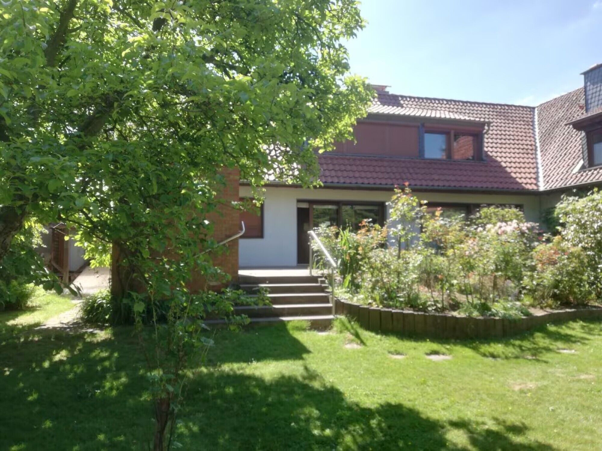 A house with a red-tiled roof, a front porch, and a garden with various plants.